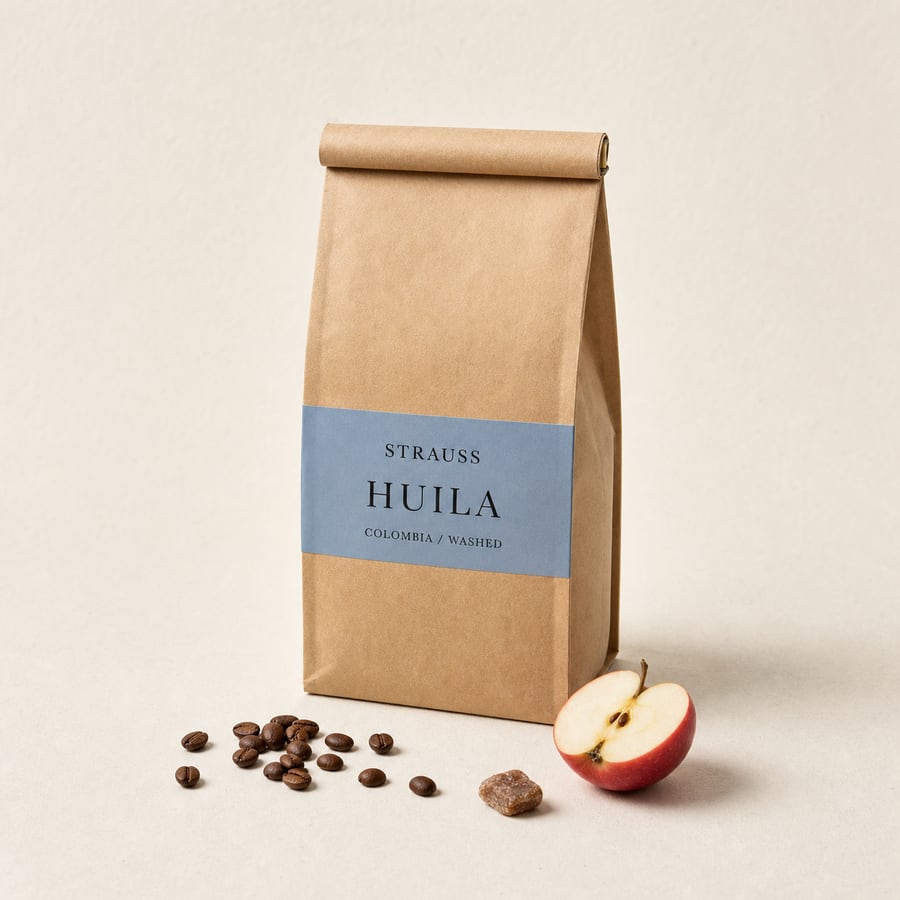 Strauss Huila bag with a dusty blue label band, coffee beans and a halved red apple on warm paper.