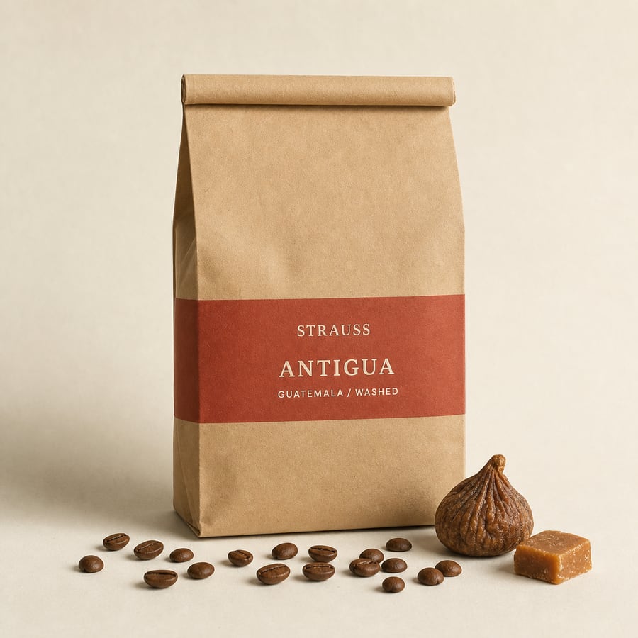 Strauss Antigua bag with a brick red label band, coffee beans, a dried fig and a small piece of toffee on warm paper.