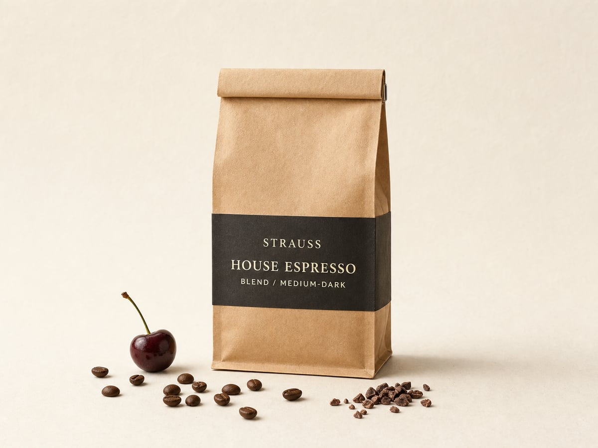 Strauss House Espresso bag with a charcoal label band, coffee beans, a dark cherry with its stalk and a pinch of cocoa nibs on warm paper.
